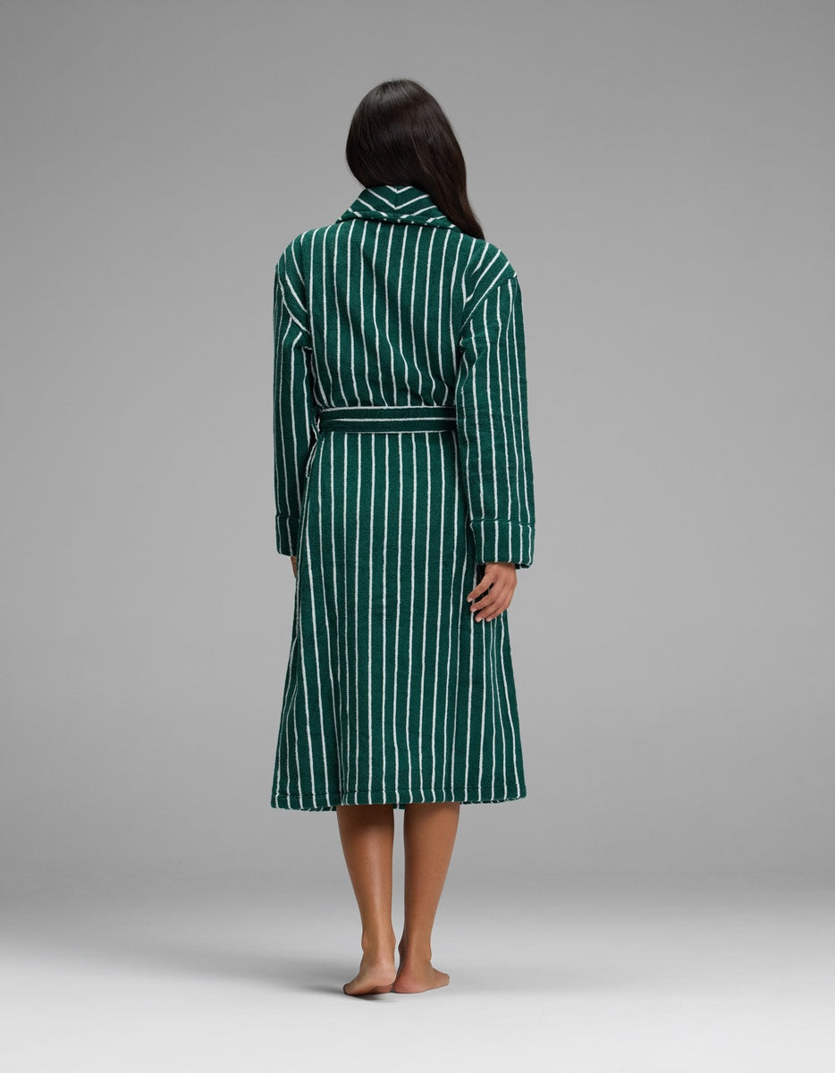 A woman stands barefoot facing away, wearing the Luxe Bath Robe by Cozy Earth—a green and white striped, belted robe—against a plain gray background.