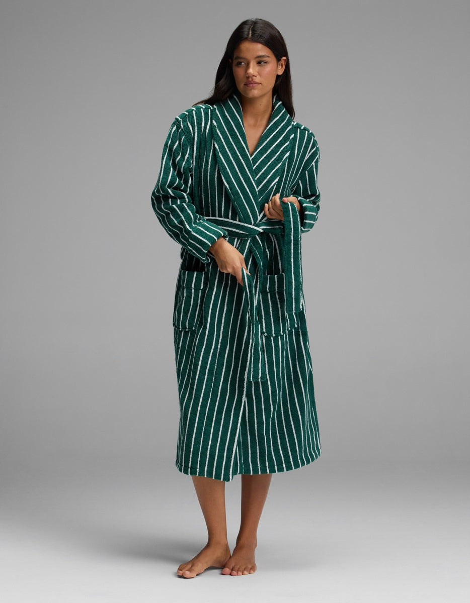 A woman stands barefoot against a plain gray background, wearing the Cozy Earth Luxe Bath Robe in green and white stripes and holding its waist tie.