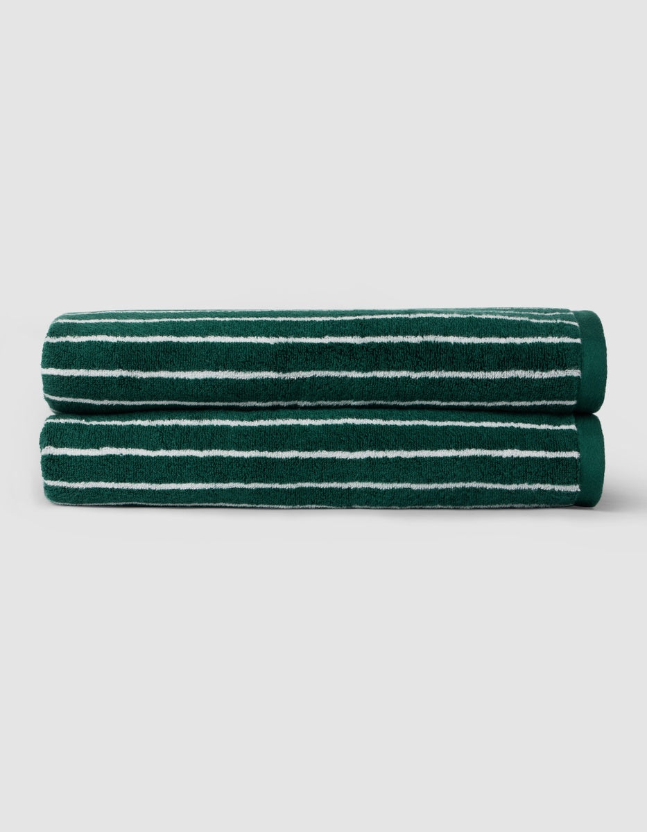 Two Luxe Bath Sheets by Cozy Earth in dark green with thin white stripes are neatly folded and stacked against a plain light gray background.