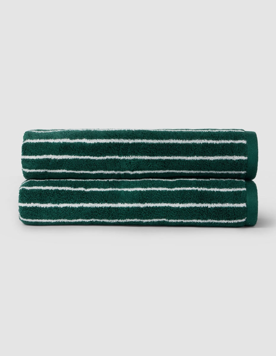 Two Luxe Bath Towels by Cozy Earth, dark green with white horizontal stripes, are neatly rolled and stacked on a light gray background.