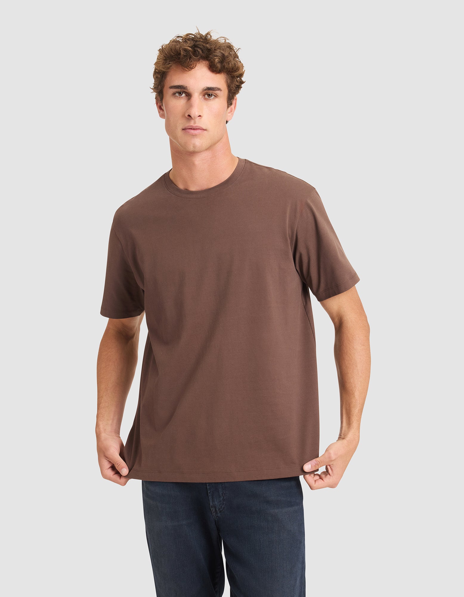 A young man with curly brown hair wears the Cozy Earth Men's All Day Tee, a loose-fitting brown short-sleeve shirt, paired with dark blue jeans against a light gray background. |Color:Cacao