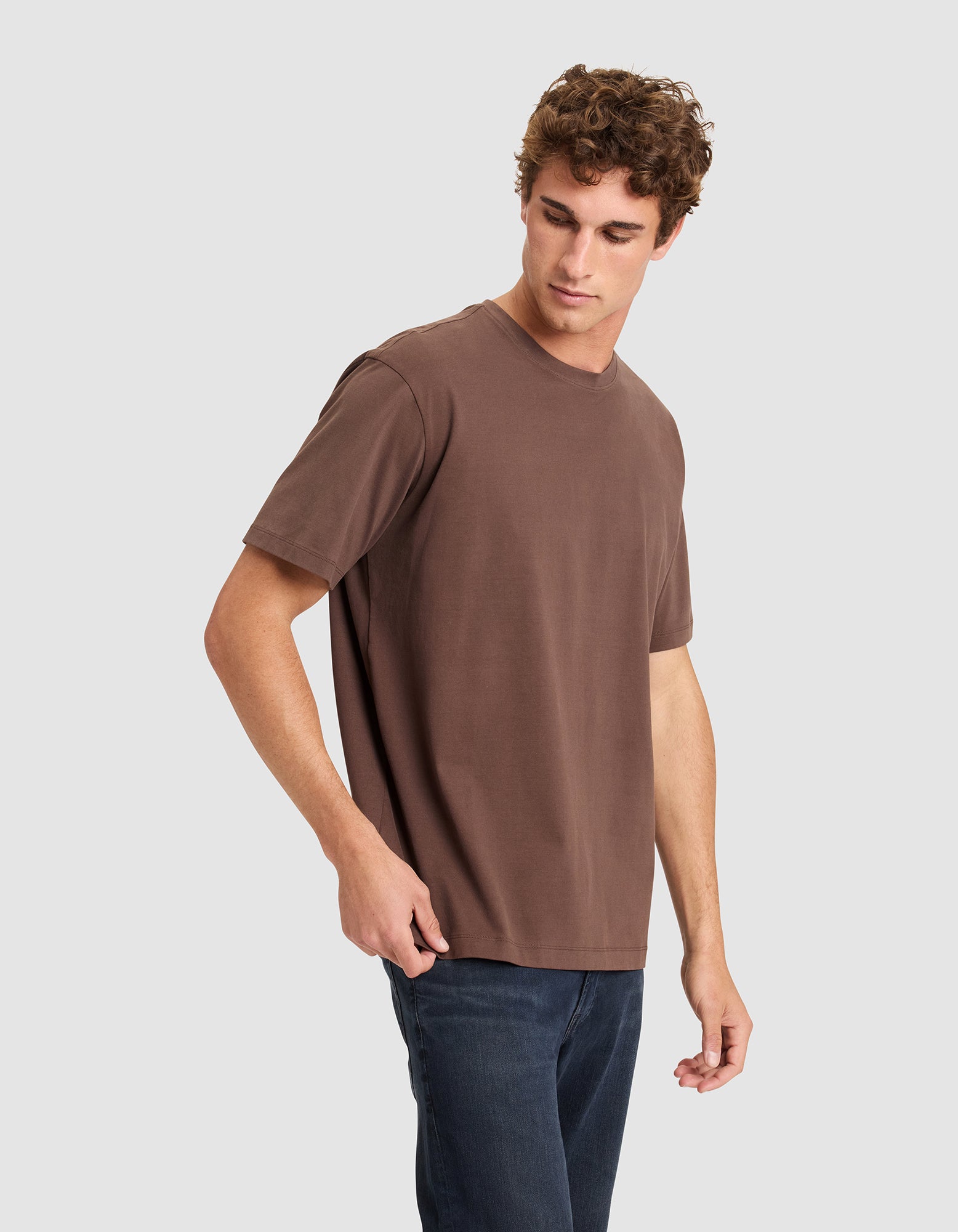 A young man with curly hair models the Cozy Earth Men's All Day Tee in brown and dark jeans, standing against a plain light gray background and looking down to his right.|Color:Cacao