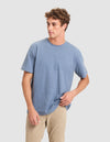 A young man with curly brown hair models the Cozy Earth Men's All Day Tee in light blue, paired with beige pants. He stands against a plain light gray background, gazing left and holding the hem of his shirt. |Color:Deep Blue Heather