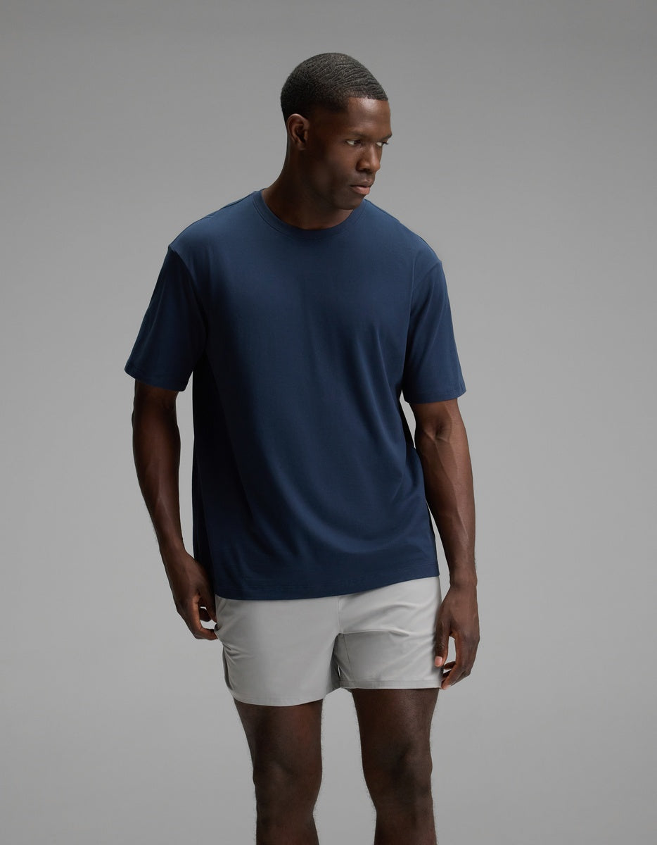 A man wearing the Cozy Earth Men’s All Day Tee in Eclipse stands against a plain gray background, looking to his left with a neutral expression.