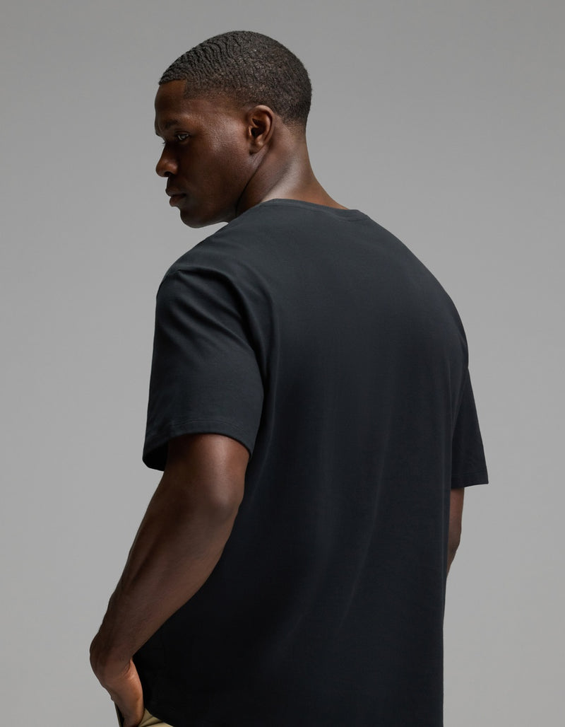 A man models the Cozy Earth Men’s All Day Tee in black, standing with his back to the camera and glancing over his left shoulder against a simple grey background.