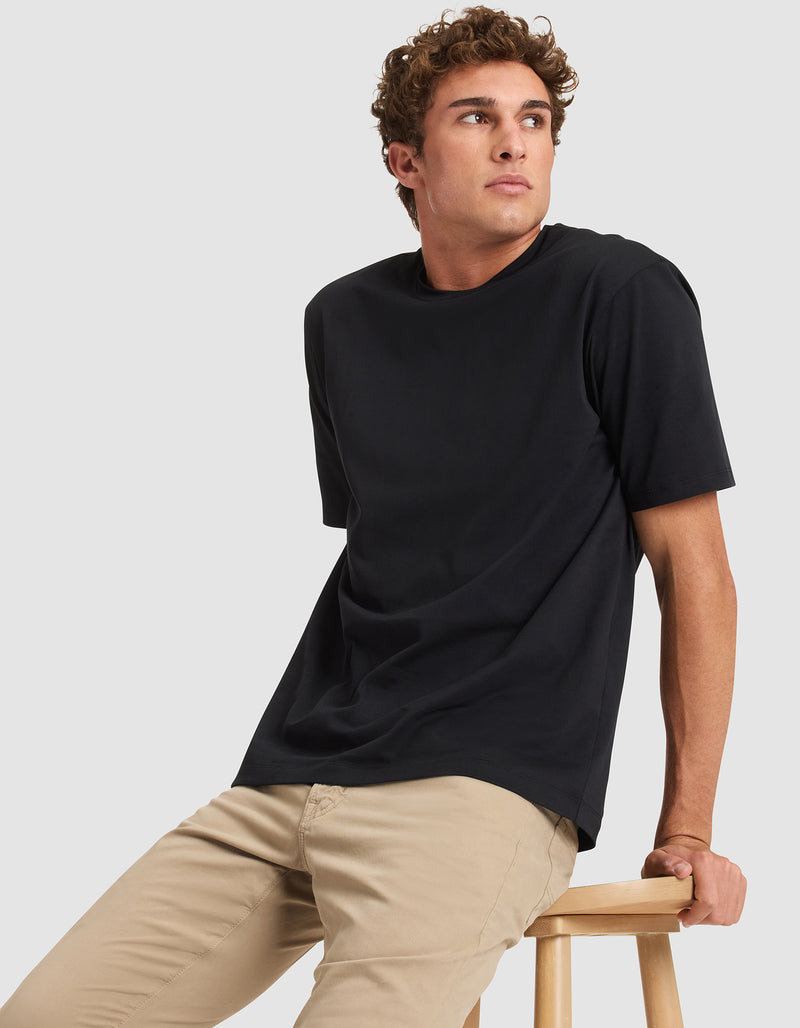 A young man with curly brown hair, wearing the Cozy Earth Men's All Day Tee in black and beige pants, sits on a wooden stool against a light gray background, looking off to the side.
