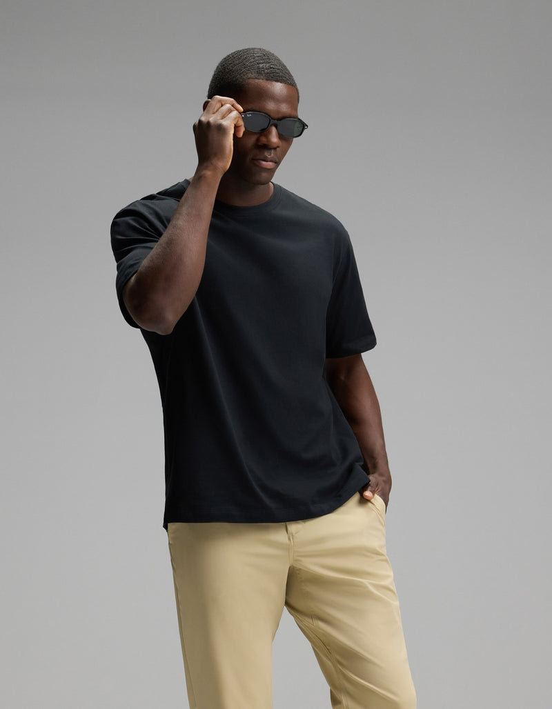 A man wearing the Cozy Earth Men’s All Day Tee in black and beige pants stands against a plain gray background, with one hand adjusting his sunglasses and the other in his pocket.