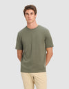 A young man with short curly brown hair models the Cozy Earth Men's All Day Tee in olive green, paired with beige pants, standing against a plain light gray background with his hands behind his back.|Color:Olive