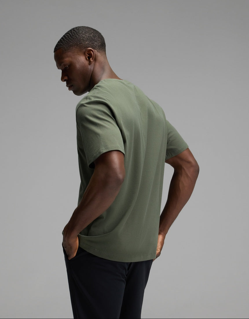 A man is seen from behind, wearing the Cozy Earth Men’s All Day Tee in olive green paired with black pants, standing against a plain gray background and looking slightly downward.
