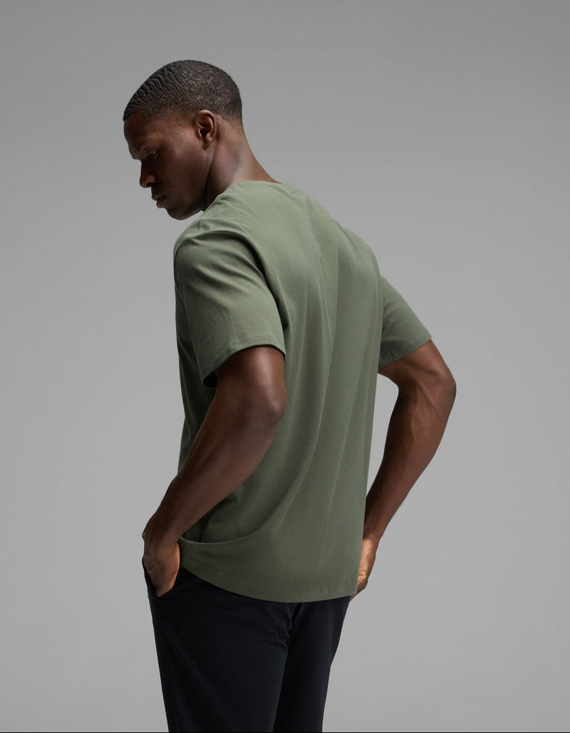 A man is seen from behind, wearing the Cozy Earth Men’s All Day Tee in olive green paired with black pants, standing against a plain gray background and looking slightly downward.