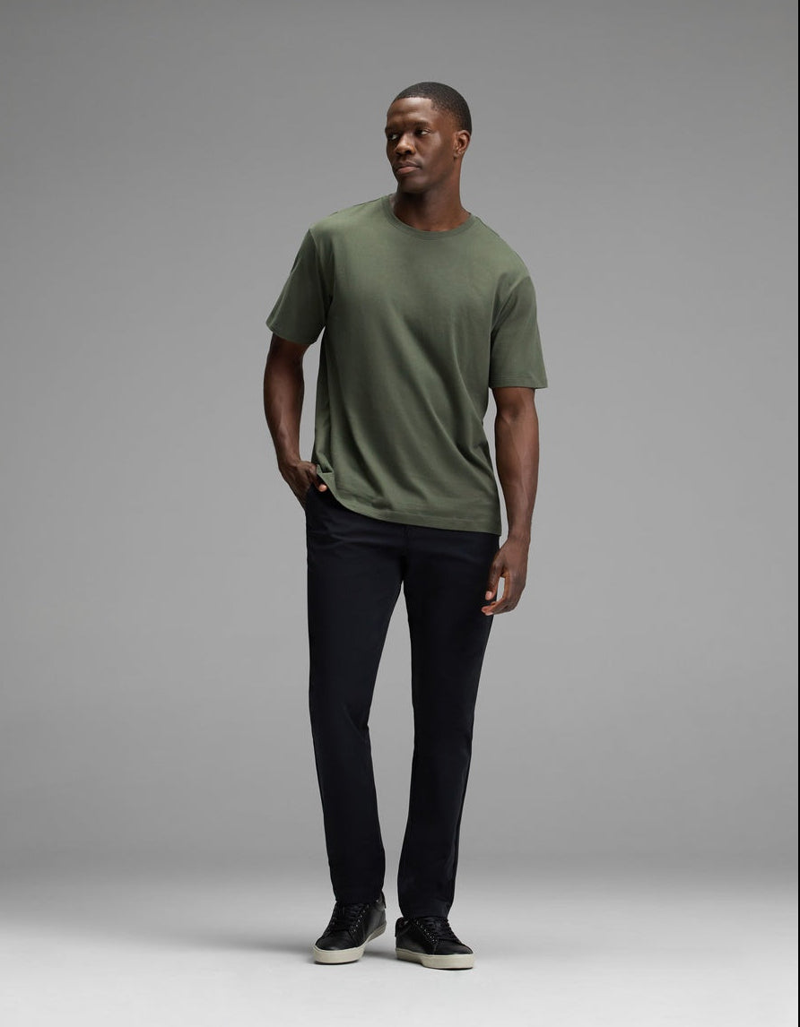 A man stands against a gray background, wearing the Cozy Earth Men’s All Day Tee in green, paired with black pants and black sneakers. He has one hand in his pocket and looks off to the side.