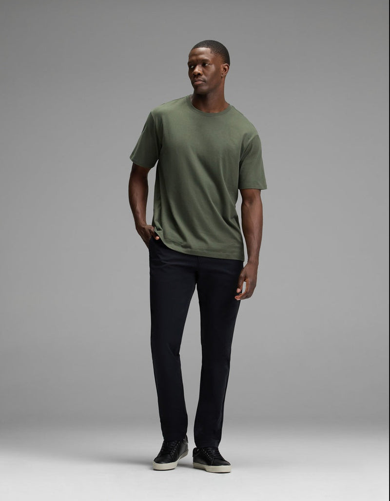 A man stands against a gray background, wearing the Cozy Earth Men’s All Day Tee in green, paired with black pants and black sneakers. He has one hand in his pocket and looks off to the side.
