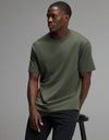 A man wearing the Cozy Earth Men’s All Day Tee in olive and black pants sits on a black stool against a plain gray background, looking to his left with a neutral expression.|Color:Olive