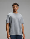 A man with short curly hair, wearing the Cozy Earth Men’s All Day Tee in Stone Heather and dark pants, stands against a plain gray background, looking directly at the camera with a neutral expression.|Color:Stone Heather