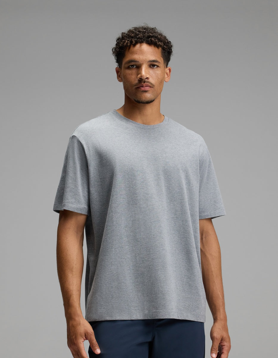 A man with short curly hair, wearing the Cozy Earth Men’s All Day Tee in Stone Heather and dark pants, stands against a plain gray background, looking directly at the camera with a neutral expression.|Color:Stone Heather