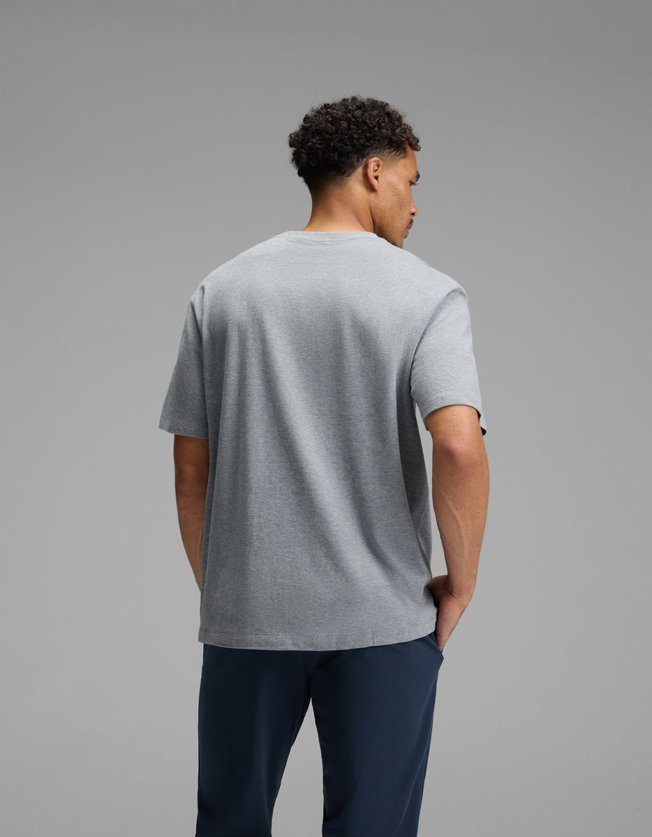 A man with short curly hair stands facing away from the camera, wearing the Cozy Earth Men’s All Day Tee in gray and dark blue pants against a plain gray background.|Color:Stone Heather