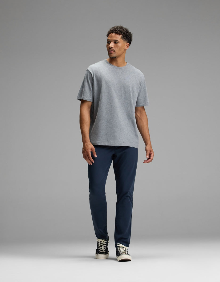 A man stands against a plain gray background, wearing the Cozy Earth Men’s All Day Tee in gray, paired with navy blue pants and black and white sneakers. He is looking to his left with a neutral expression.