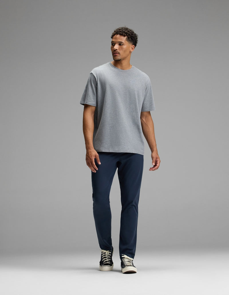 A man stands against a plain gray background, wearing the Cozy Earth Men’s All Day Tee in gray, paired with navy blue pants and black and white sneakers. He is looking to his left with a neutral expression.