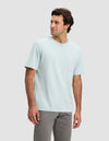 A man with dark hair models the Cozy Earth Men's All Day Tee in light blue and gray pants, standing against a plain light gray background, looking slightly to his left and smiling.|Color:Tide