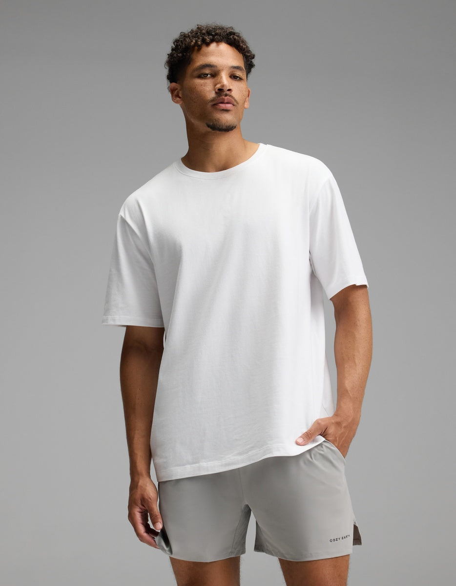 A man stands against a gray background wearing the Cozy Earth Men’s All Day Tee in white and light gray shorts, with one hand in his pocket and a neutral expression.