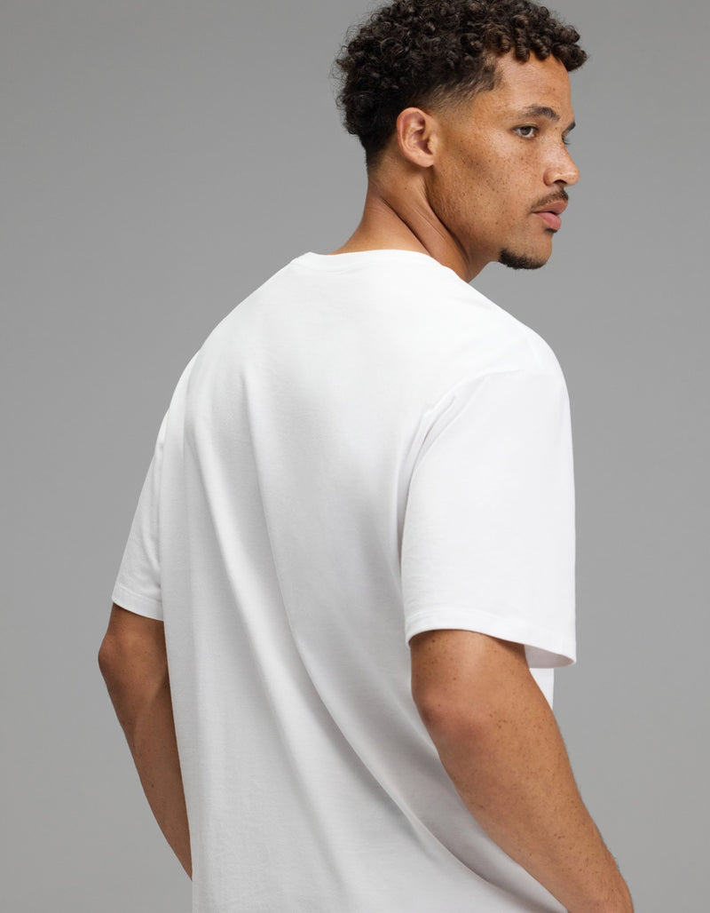 A man with short curly hair and a trimmed beard, wearing the Cozy Earth Men’s All Day Tee in plain white, turns to look over his right shoulder against a neutral gray background.