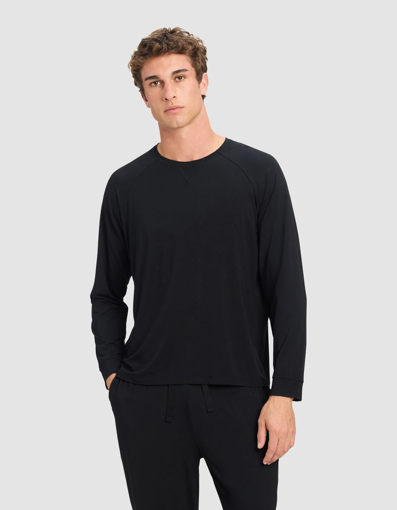 A young man with short, curly brown hair models the Cozy Earth Men's Bamboo Stretch-Knit Long Sleeve in black, paired with matching pants. He stands against a light gray background with a neutral expression and one hand in his pocket.