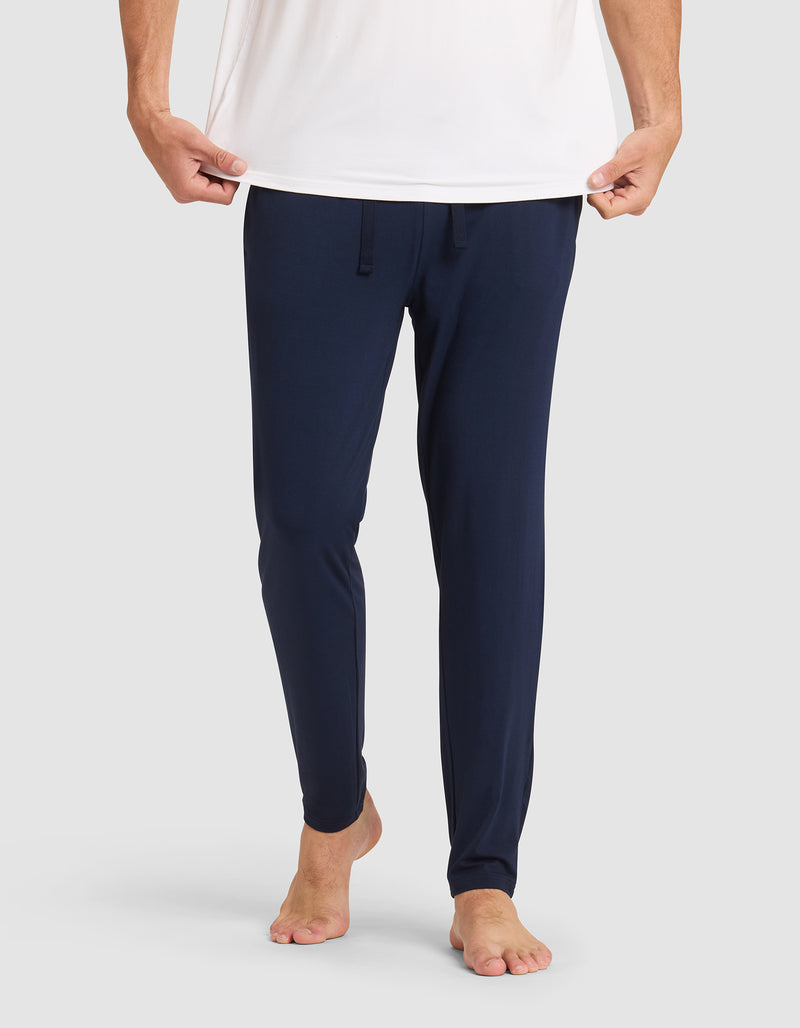 Only the lower half of a person is shown against a plain light background, barefoot and wearing Cozy Earth's Men's Bamboo Stretch-Knit Pajama Pant in navy blue with a drawstring waist and a white shirt, holding the shirt's hem.