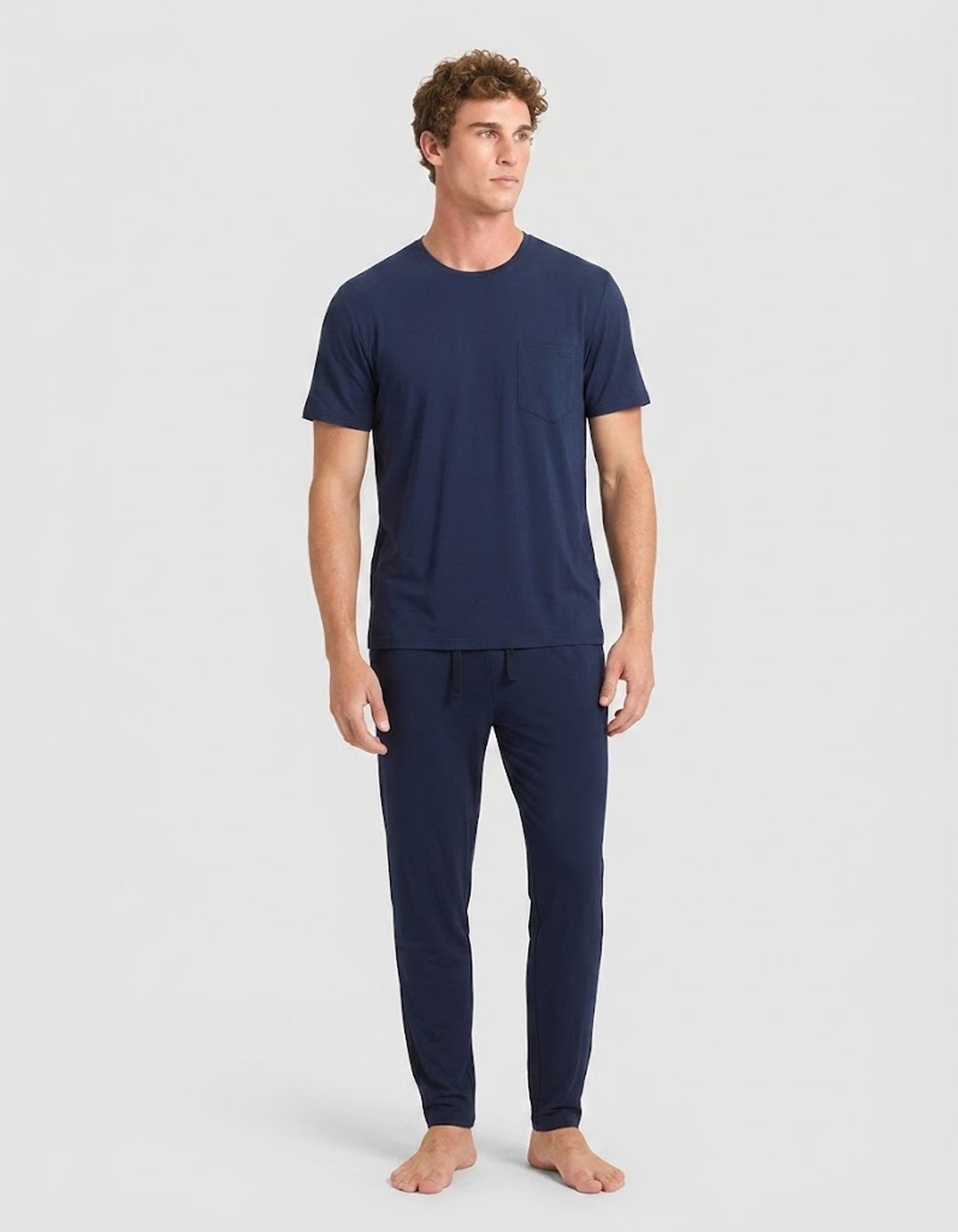 A man stands barefoot against a light gray background, wearing Cozy Earth’s Men's Bamboo Stretch-Knit Lounge Tee & Pant Pajama Set in navy.|Color:Navy