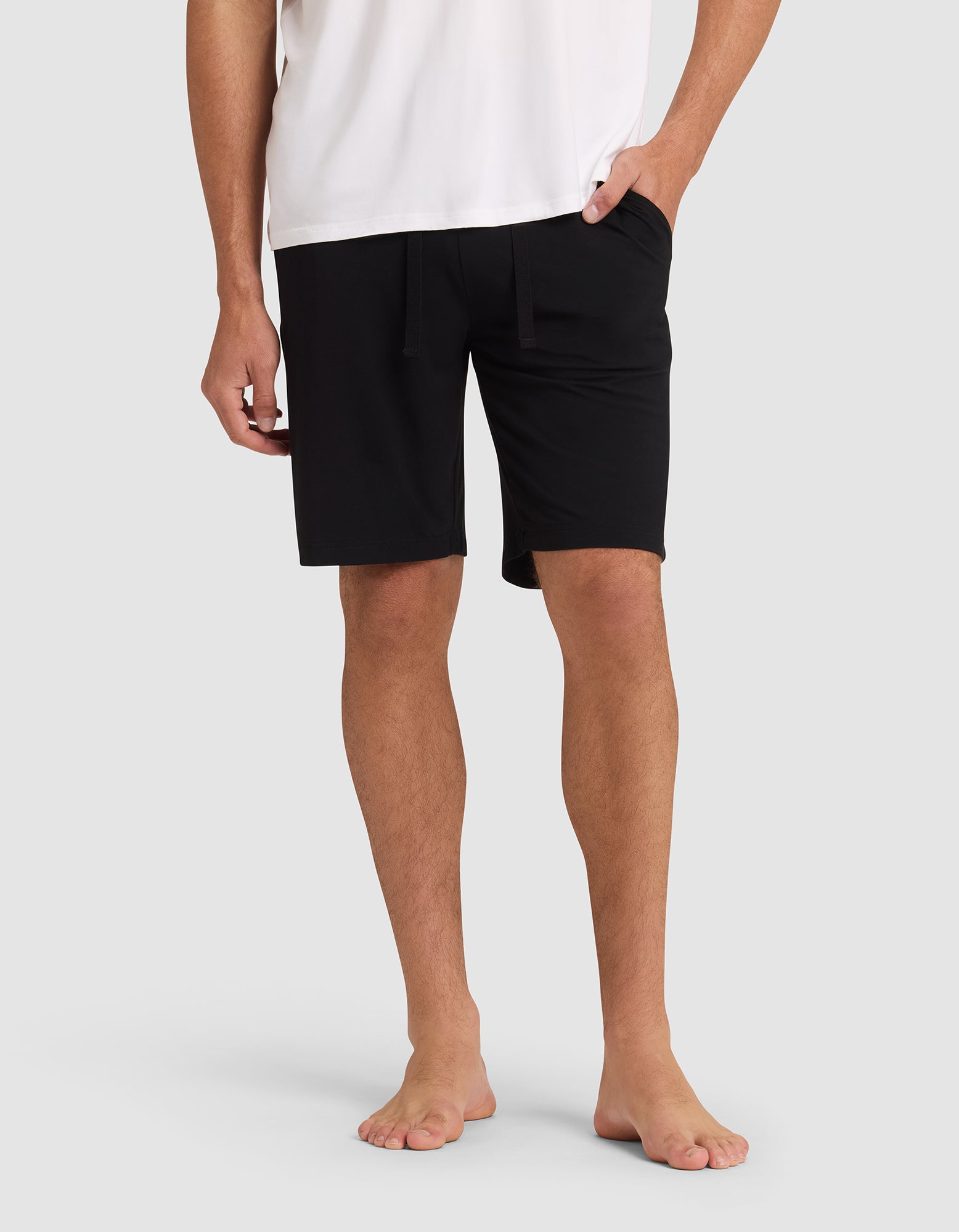 A person stands barefoot against a light gray background, wearing Cozy Earth Men's Bamboo Stretch-Knit Pajama Shorts in black with a white T-shirt. Their hands are relaxed—one in a pocket, the other by their side.