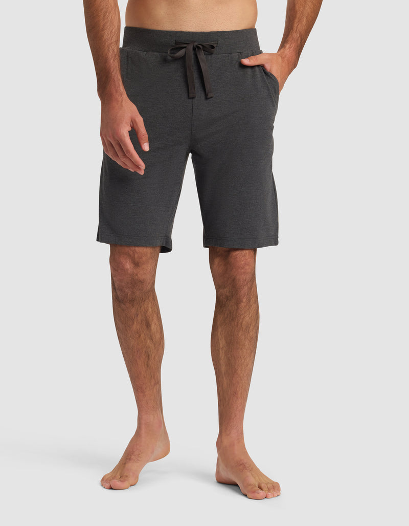 A man models Cozy Earth Men's Bamboo Stretch-Knit Pajama Short in dark gray, barefoot against a plain white background, with one hand by his side and the other in his pocket.