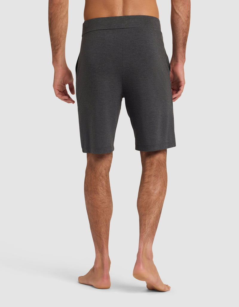 A man stands barefoot facing away, wearing Cozy Earth Men's Bamboo Stretch-Knit Pajama Shorts in dark gray with an elastic waistband; his arms relaxed at his sides against a plain white background.