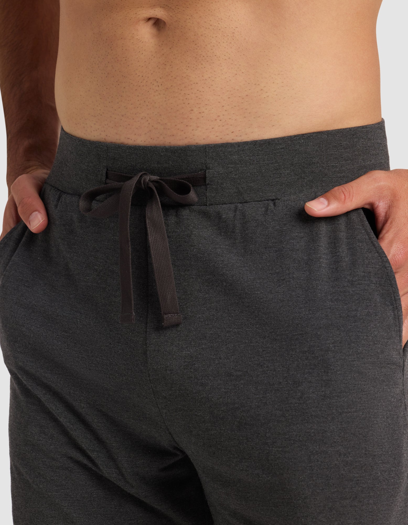 A close-up of a person wearing Cozy Earth's Men's Bamboo Stretch-Knit Pajama Short in dark gray, with hands in the pockets. The shirtless model is shown from waist to upper thighs, highlighting the comfortable stretch-knit fabric.