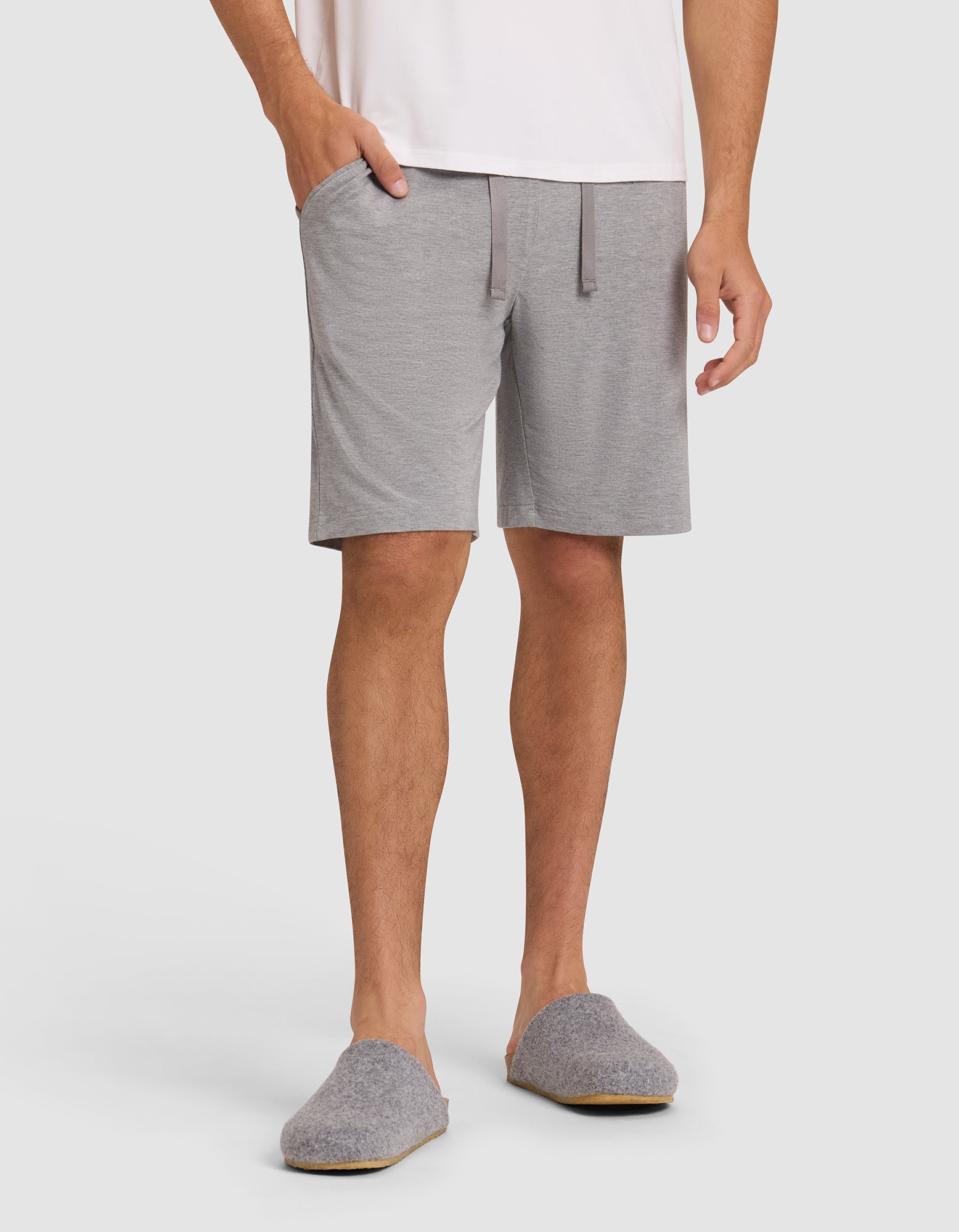 Only the lower half of a person is visible as they wear Cozy Earth's Men's Bamboo Stretch-Knit Pajama Short in light gray with pockets, paired with gray slippers and a white shirt, standing against a plain background. 
