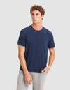 A young man with curly hair models the Cozy Earth Men's Bamboo Stretch-Knit Lounge Tee in navy blue with short sleeves and pocket, paired with light gray pants, standing against a plain light gray background.  |Color:Navy