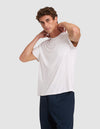 A young man stands against a plain light gray background, wearing Cozy Earth’s Men’s Bamboo Stretch-Knit Lounge Tee in white with a chest pocket and dark pants, his hands resting on the back of his neck.  |Color:White