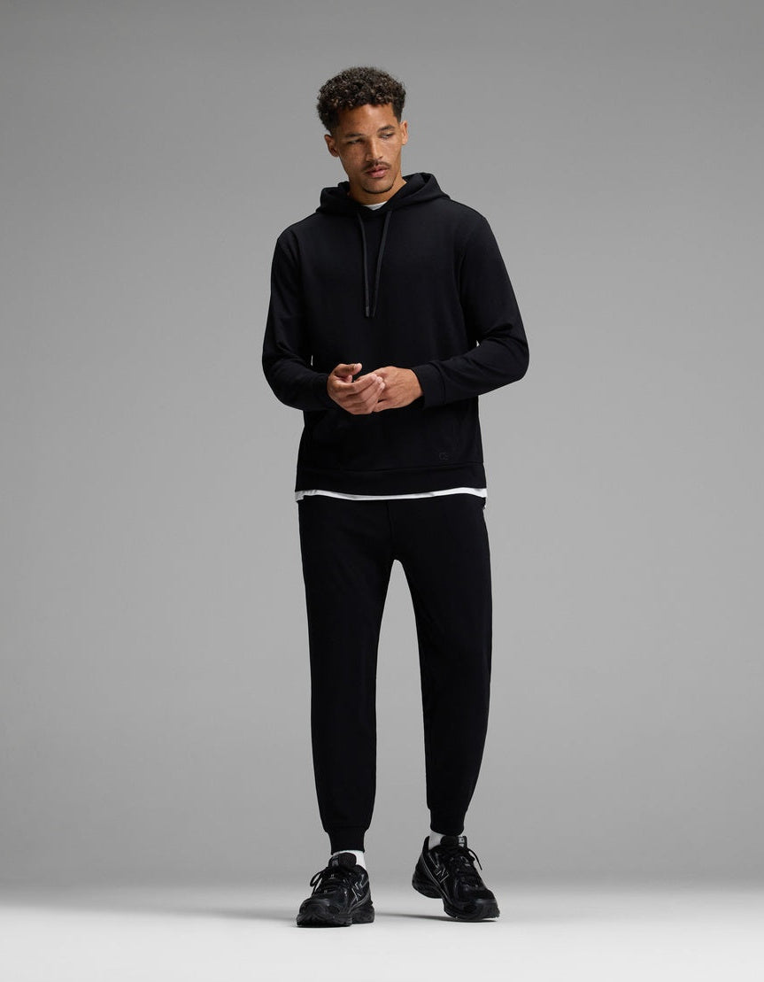 A man stands against a gray background, wearing the Cozy Earth Men’s Brushed Bamboo Hoodie & Jogger Set in black, with a white T-shirt visible beneath the hoodie and black sneakers. He looks slightly to the side, hands together.|Color:Black