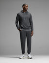 A man stands against a grey background, wearing the Cozy Earth Men’s Brushed Bamboo Hoodie & Jogger Set in Charcoal, paired with white athletic shoes. He looks to the side with his hands by his sides.|Color:Charcoal