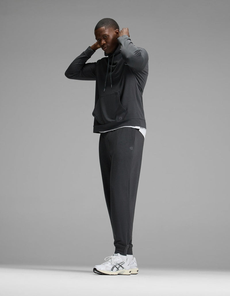 A man wearing the Cozy Earth Men’s Brushed Bamboo Hoodie & Jogger Set stands adjusting his hood, paired with white sneakers. The background is plain gray.