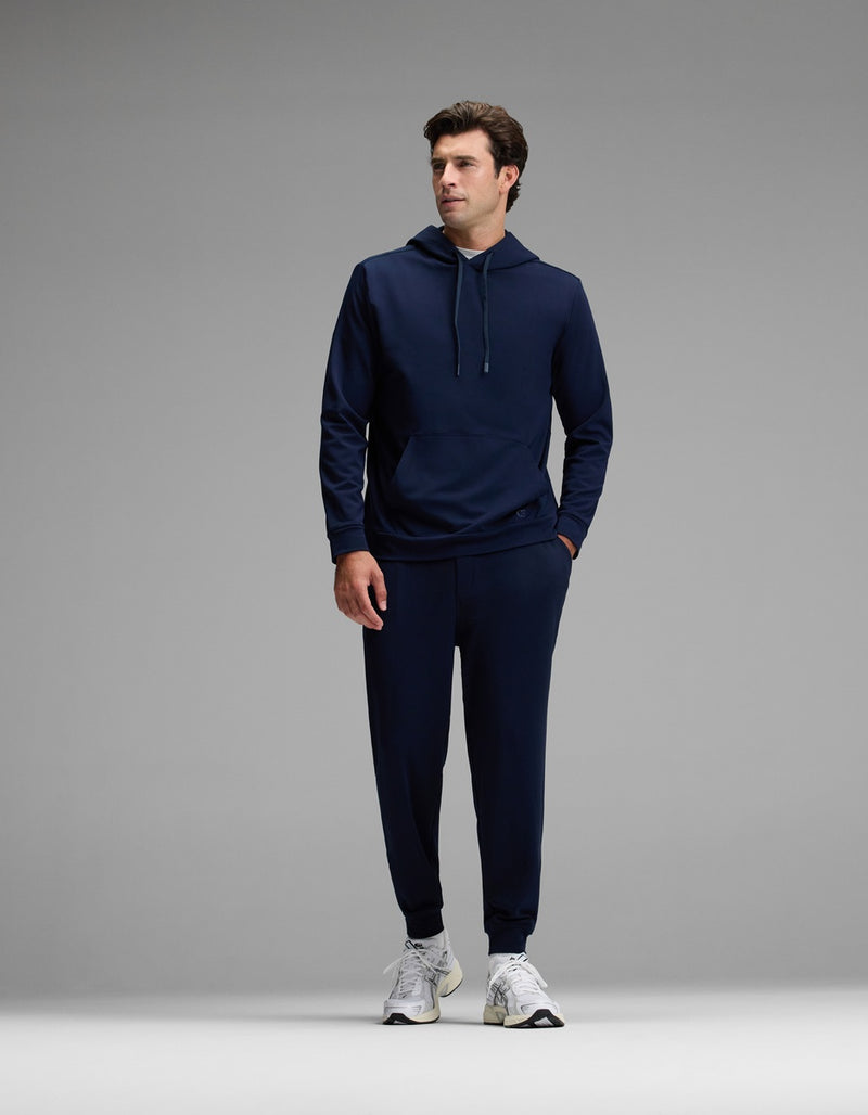 A man stands against a plain gray background, wearing the Cozy Earth Men’s Brushed Bamboo Hoodie & Jogger Set in navy blue, paired with white sneakers. He looks to his left with one hand in his pocket.