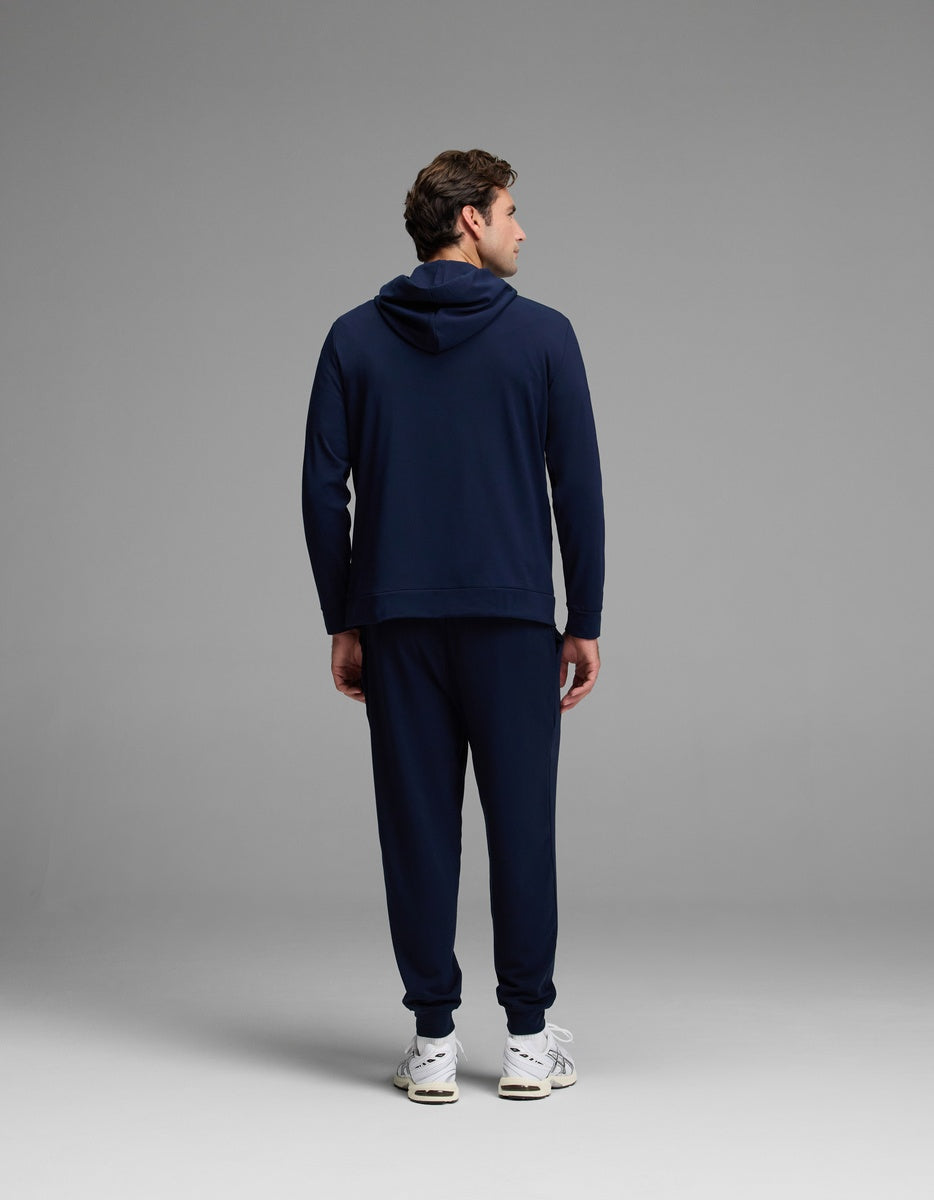 A man stands facing away from the camera in a plain, well-lit studio, wearing the Cozy Earth Men’s Brushed Bamboo Hoodie & Jogger Set with white sneakers.