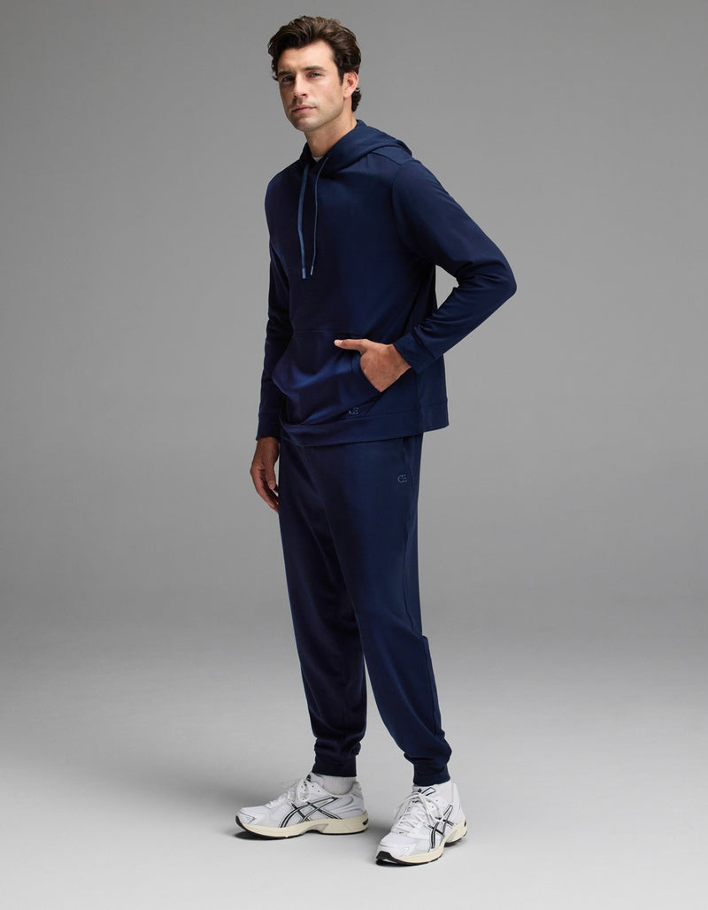 A person stands in a studio against a plain gray background wearing the Cozy Earth Men’s Brushed Bamboo Hoodie & Jogger Set in navy, paired with white sneakers.