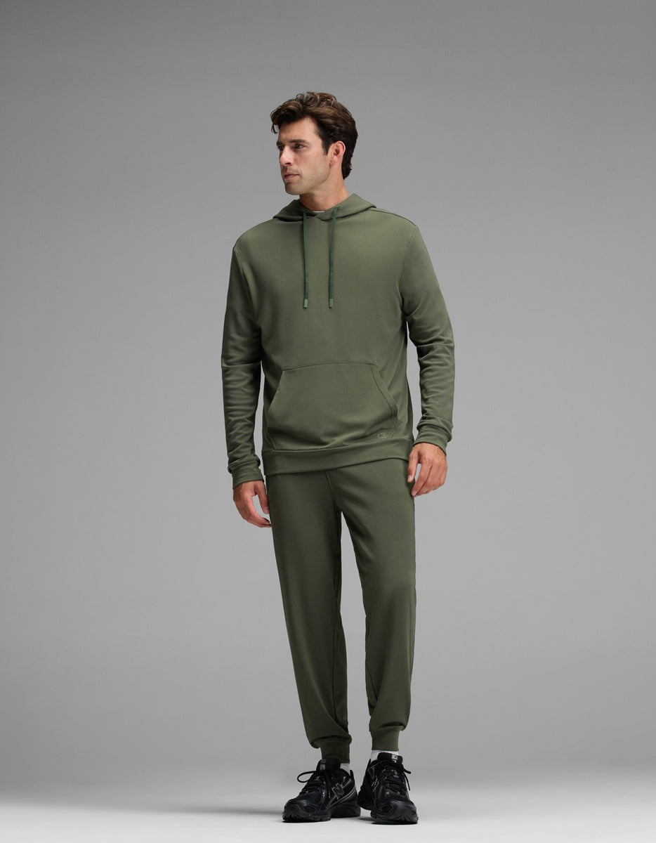 A man models the Cozy Earth Men’s Brushed Bamboo Hoodie & Jogger Set in olive, standing against a plain gray background. He looks slightly to his left with his hands relaxed at his sides, completing the look with black sneakers.|Color:Olive