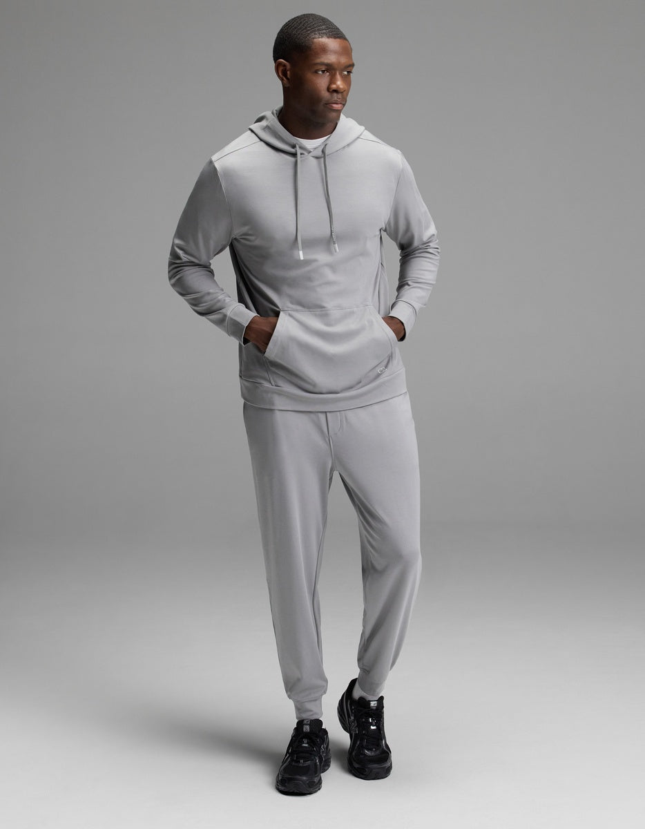 A man wearing the Cozy Earth Men’s Brushed Bamboo Hoodie & Jogger Set in Stone stands against a plain gray background, looking to his left with his hands in the hoodie pocket and black sneakers on.