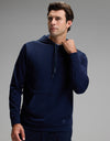 A person wearing the Cozy Earth Men’s Brushed Bamboo Hoodie in dark blue stands against a plain gray background, looking slightly to the side while touching the neckline of the hoodie with one hand.|Color:Navy