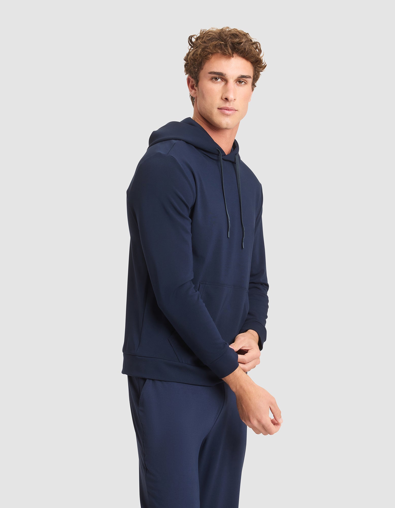 A young man with curly hair models the Cozy Earth Men's Brushed Bamboo Hoodie in navy blue, paired with matching pants, standing against a light gray background and looking at the camera with a neutral expression. 