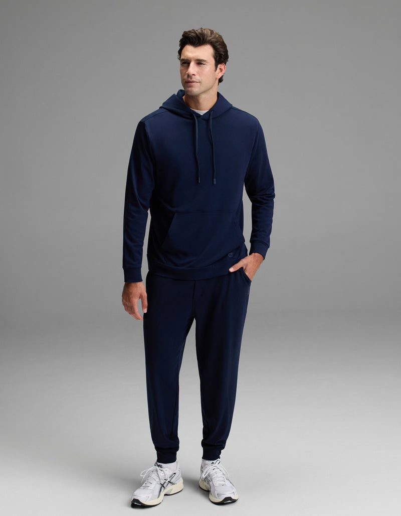 A man stands against a gray background wearing the Cozy Earth Men’s Brushed Bamboo Hoodie in navy blue, paired with matching jogger pants and white sneakers. He has one hand in his pocket and looks slightly to the side.