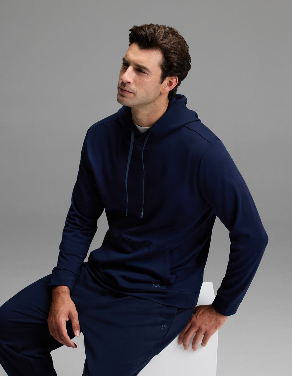 A man with short brown hair, dressed in the Cozy Earth Men’s Brushed Bamboo Hoodie and matching pants in navy blue, sits on a white block against a plain gray background.
