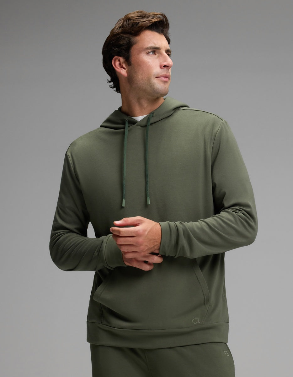 A man stands wearing the Cozy Earth Men’s Brushed Bamboo Hoodie in olive green, paired with matching pants. He looks to his left with his hands clasped, set against a plain light gray background.