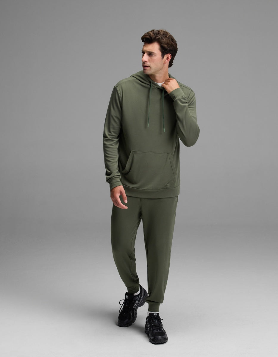 A man wears the Cozy Earth Men’s Brushed Bamboo Hoodie in olive green, paired with matching joggers, as he stands against a plain gray background, gazing to his left with one hand touching his neck and the other resting near his pocket.