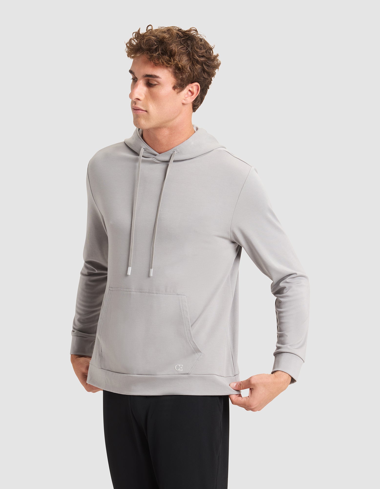 A young man stands against a plain light gray background, wearing Cozy Earth's Men's Brushed Bamboo Hoodie in light gray and black pants, looking to the side with his hands pulling at the hem of the hoodie. |Color:Stone